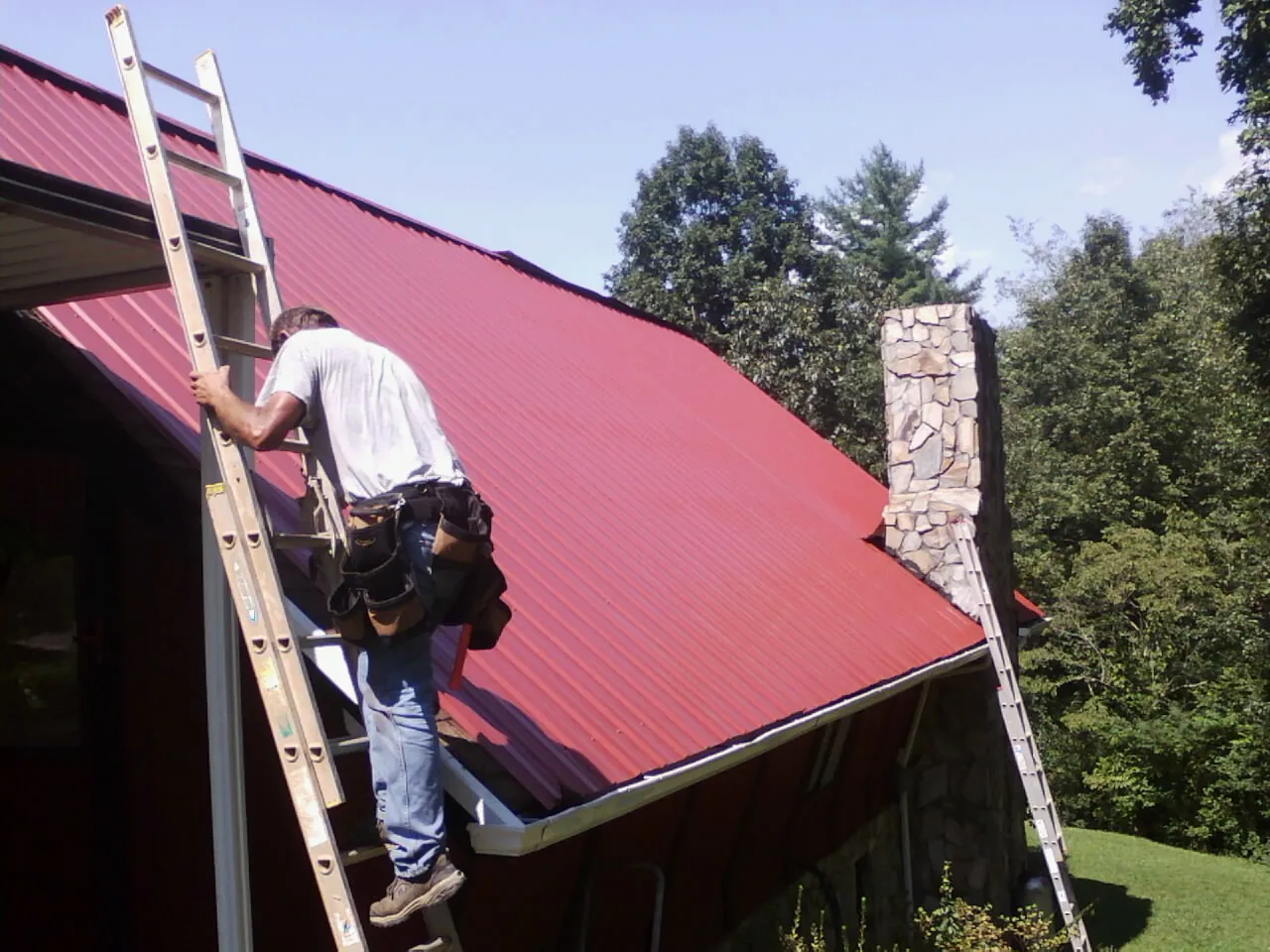 Roofer on a ladder beside a red metal roof and stone chimney — Soffit & Fascia Repair by Roofing Brothers in Hanceville